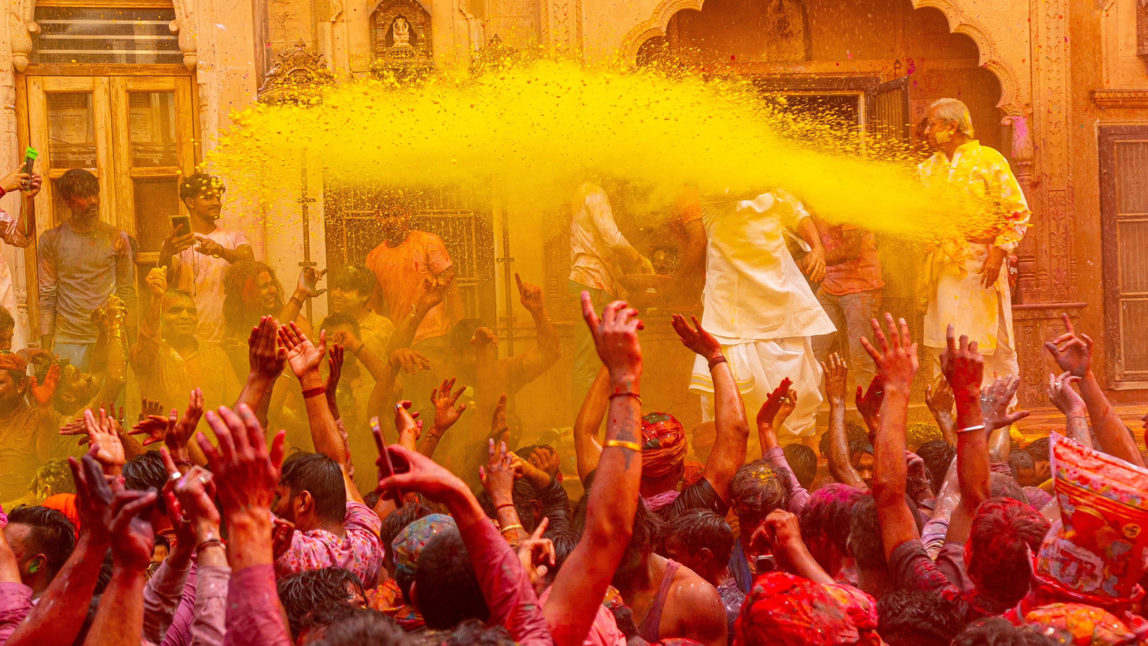 A crowd in white clothes covered in pink and yellow gulal powder throwing colours into the air in a Vrindavan temple courtyard