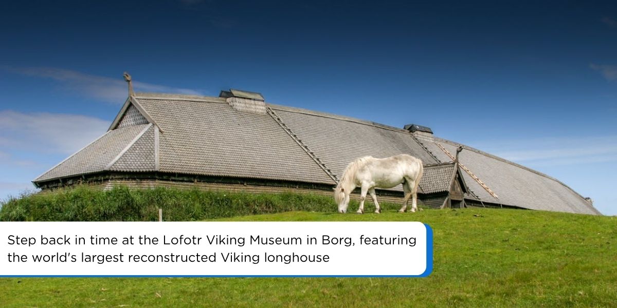 A white horse grazes on a green grassy hill in front of the historic wooden Viking longhouse at the Lofotr Viking Museum in Norway, under a blue sky with dark clouds.