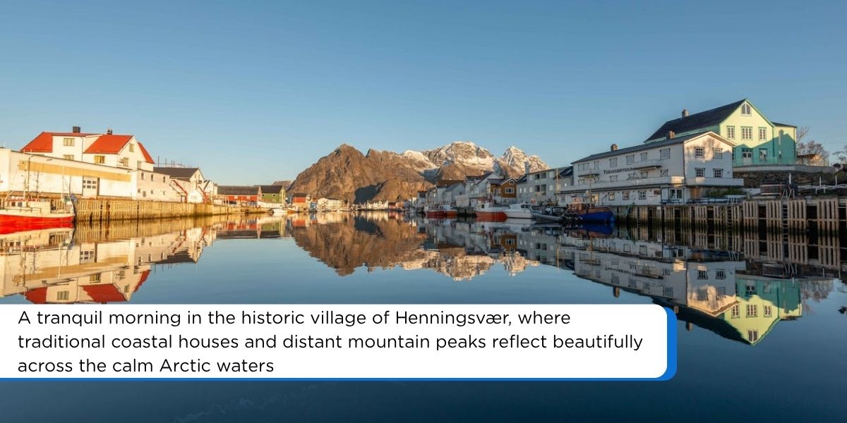 Colorful waterfront buildings and snow-capped mountains in the background create a perfect, clear reflection on the mirror-like calm water of the fjord under a clear blue sky.