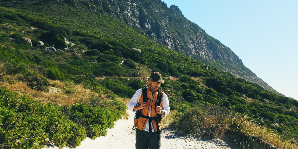 Male traveler hiking a sunlit trail with a small backpack and outdoor gear.
